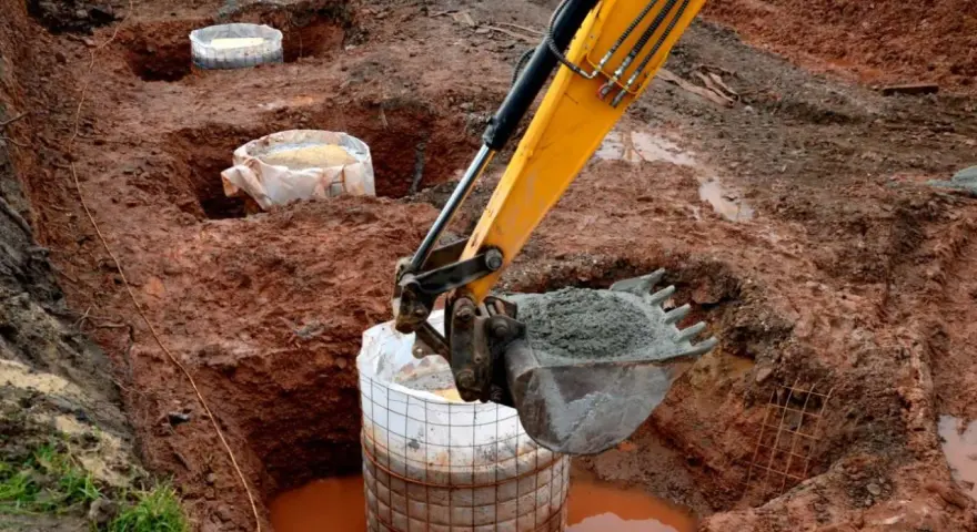 Koparka zasypuje betonem fundament palowy w wykopie. W tle widać kolejne przygotowane do zalania pale.