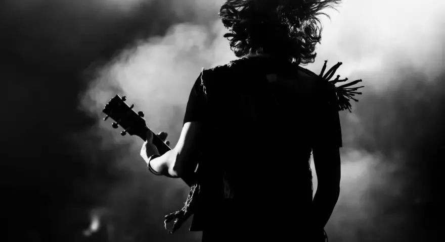 Muzyk z rozwianymi włosami gra na gitarze, w dymie i świetle sceny. Energia gatunku rockowego w czerni i bieli.