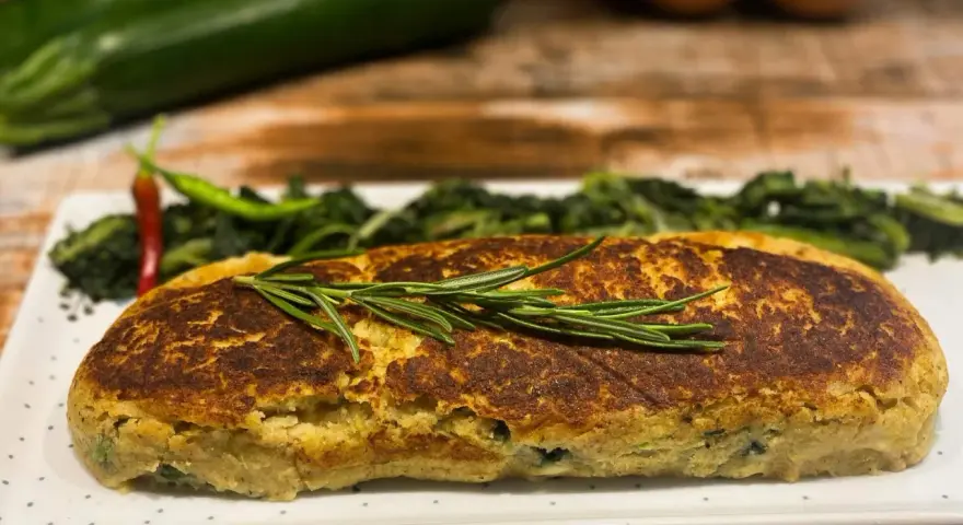 Polpettone di verdure dorato, guarnito con rosmarino, servito con contorno di verdure verdi e peperoncino.