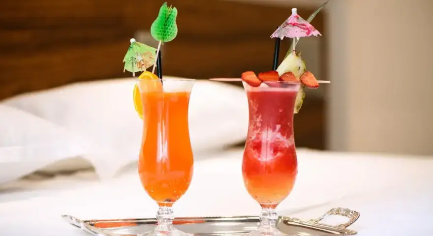 Dwa orzeźwiające drinki: jeden pomarańczowy z pomarańczą, drugi czerwony z truskawkami i ananasem, oba z parasolkami. Idealne na wakacje, może z dodatkiem triple sec.