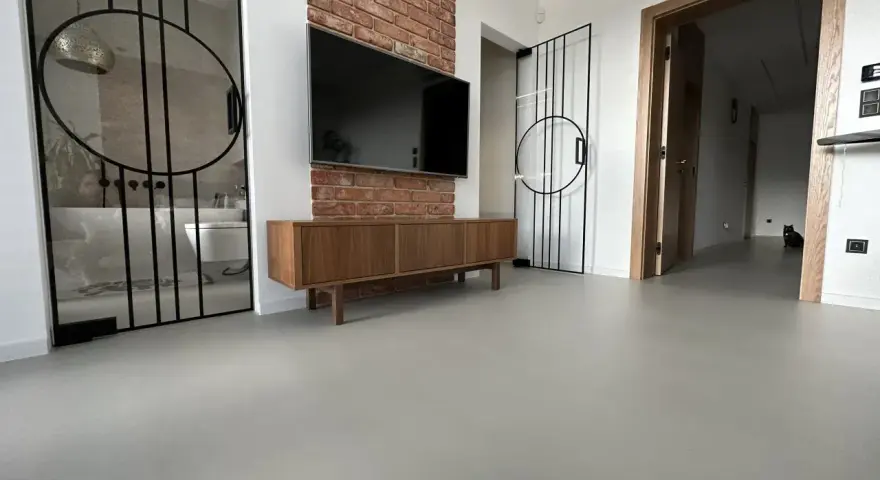 Nowoczesne wnętrze z betonową podłogą, co to jest mikrocement, która nadaje mu surowego charakteru. W tle widać łazienkę i salon.