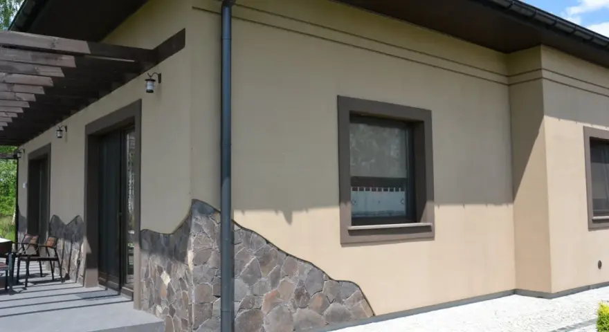 Elewacja domu w kolorze kawa z mlekiem, ozdobiona kamiennym cokołem. Widoczne okna i drzwi tarasowe.