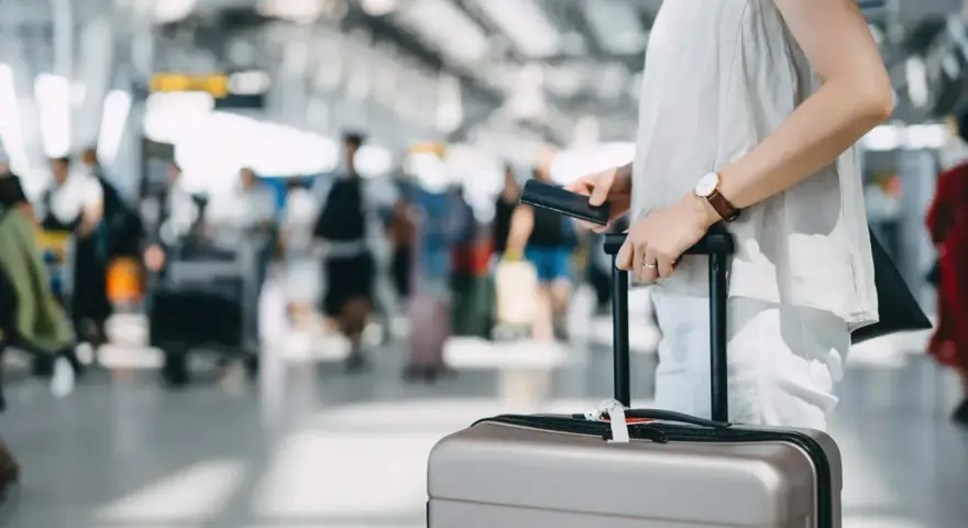 A traveler holds a passport and suitcase, mindful of carry-on size limits in a busy airport.