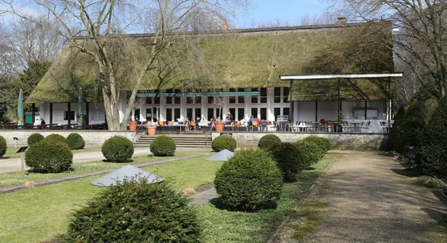 Das Teehaus im Englischen Garten Berlin mit seinem Reetdach und der Terrasse, auf der Gäste sitzen, ist von gepflegten Rasenflächen und Bäumen umgeben.