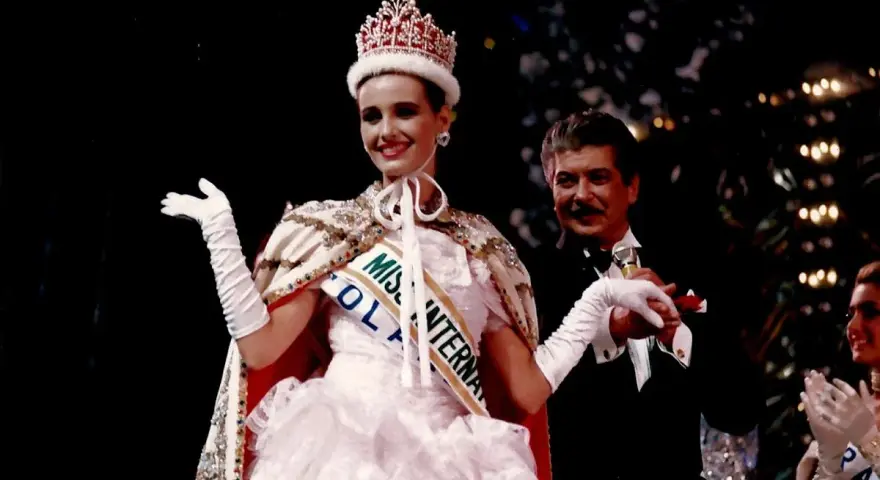 Uśmiechnięta Miss International w koronie i białej sukni, z szarfą "Miss Polonia". Obok niej mężczyzna w smokingu.