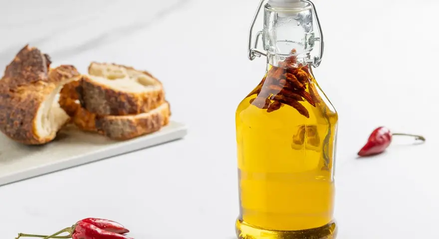 Olio al peperoncino in bottiglia, con peperoncini rossi e pane rustico a fette sullo sfondo.