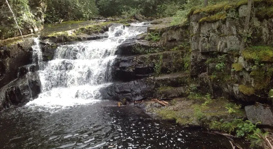 Vattenfall i en skogsmiljö nära Lillehammer, Norge. Mossbeklädda klippor och mörkt vatten.