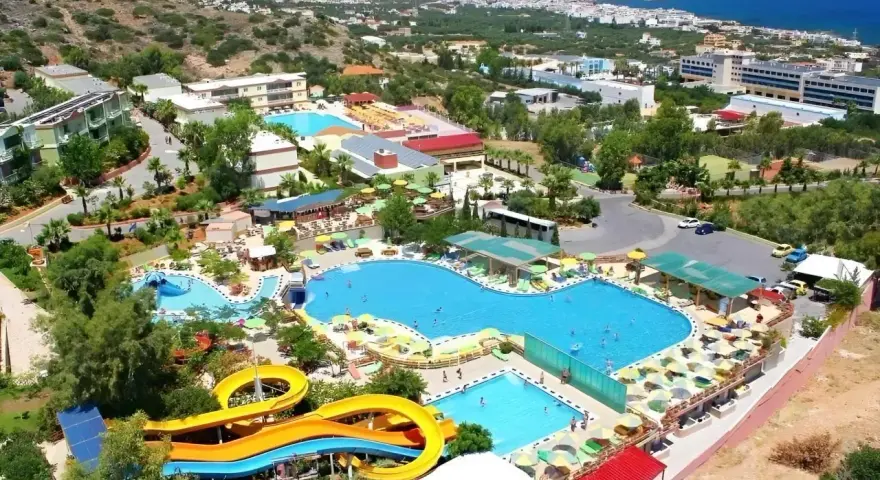 Widok z lotu ptaka na kompleks basenów z atrakcjami wodnymi i leżakami. Wiele osób korzysta z uroków słońca i wody. W tle widać budynki hotelowe i morze. Aqua Sun Village opinie potwierdzają, że to idealne miejsce na wakacje.
