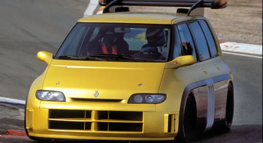 Żółty Renault Espace F1 z kierowcą w kasku na torze wyścigowym. Potężny spojler dachowy i agresywny wygląd.