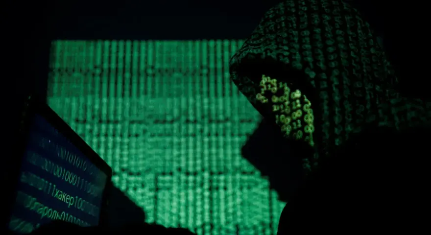 A hooded figure works on a laptop, bathed in the green glow of binary code. This scene evokes the shadowy world of China cyber espionage.