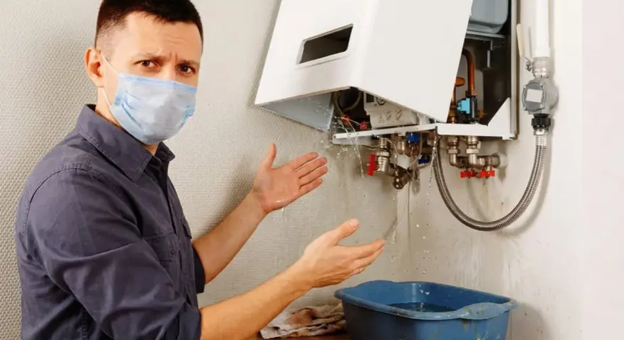 Uomo con mascherina mostra con le mani la caldaia a condensazione che perde acqua dal tubo di scarico, raccogliendola in una bacinella.