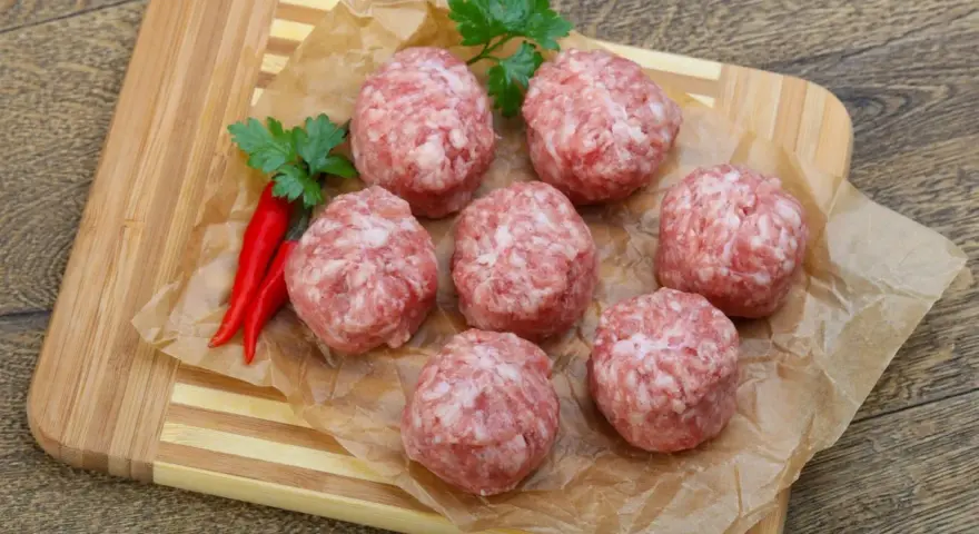 Surowe kotlety mielone keto, przyprawione papryczkami chili i natką pietruszki, leżą na papierze do pieczenia na desce.