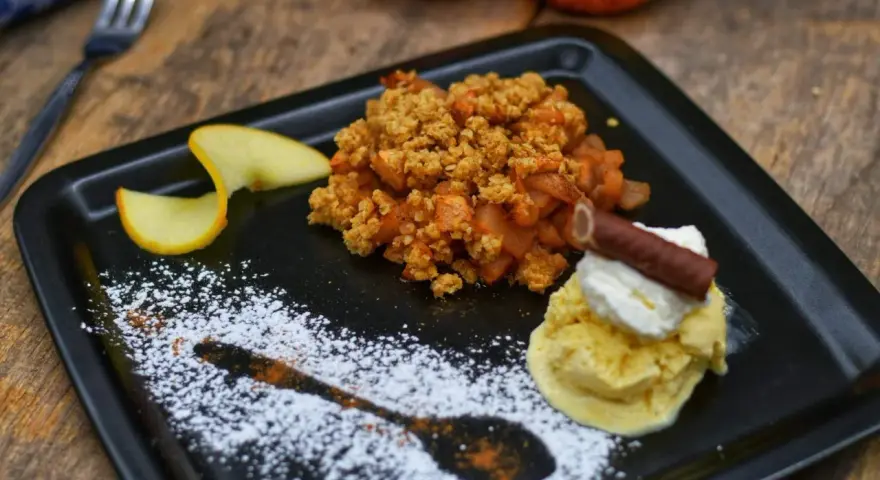 Ein köstlicher Apfel Crumble mit Haferflocken, serviert mit Vanilleeis, Sahne und einem Schokostäbchen.