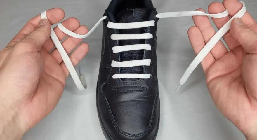 Mani che mostrano come mettere i lacci alle scarpe senza fiocco, con un laccio bianco che attraversa gli occhielli di una scarpa nera.