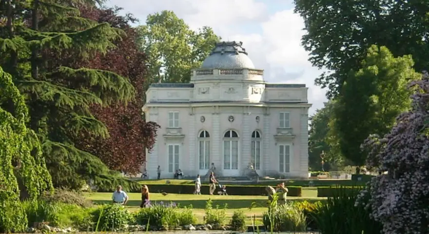 Un elegante edificio blanco con cúpula se alza en un exuberante jardin des plantes, con un estanque en primer plano y gente paseando.