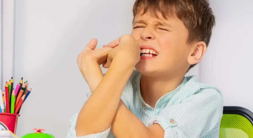 Junge mit zusammengepressten Augen und Händen vor dem Gesicht, ein Zeichen dafür, wie mit Tics bei Kindern umgehen. Buntstifte und eine Klingel liegen auf dem Tisch.