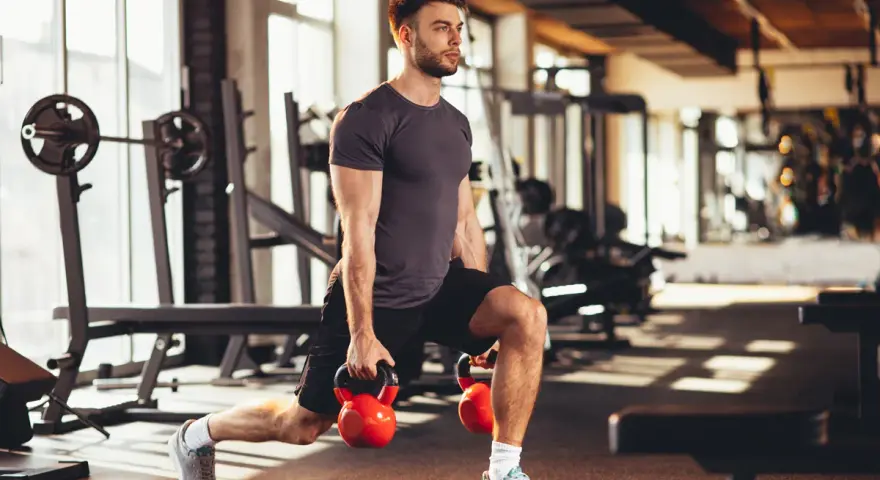 Mężczyzna wykonuje wykroki z kettlebellami, realizując swój plan treningowy na siłownię.