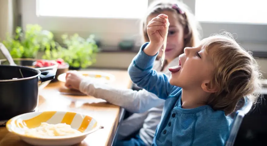 Zwei Kinder am Tisch, die schnell Nudeln essen. Der Junge nimmt eine Gabel voll Spaghetti zum Mund.
