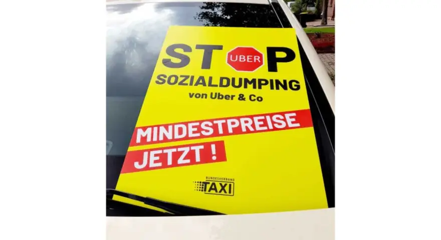 Gelbes Schild auf Taxidach: "STOP SOCIAL DUMPING von Uber & Co. MINDESTPREISE JETZT!". Fordert faire taksi fiyatları.