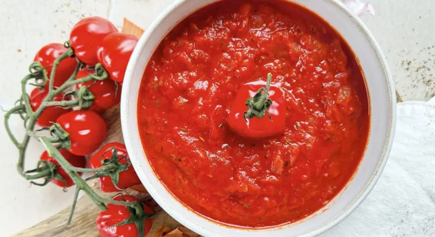 Ein Teller mit leuchtend rotem Sugo di Pomodoro, garniert mit einer einzelnen Kirschtomate. Daneben ein Bund frischer Tomaten und Zwiebeln.