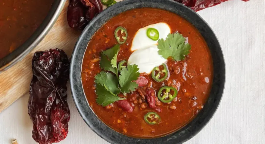 Un tazón humeante de chili con carne, adornado con crema agria, jalapeños y cilantro. Alrededor, chiles secos añaden un toque rústico.