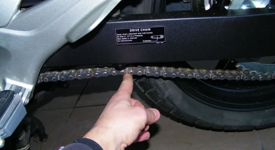 Sprawdzanie luzu łańcucha w motocyklu. Palec pokazuje, jak sprawdzić, czy łańcuch jest prawidłowo naciągnięty.