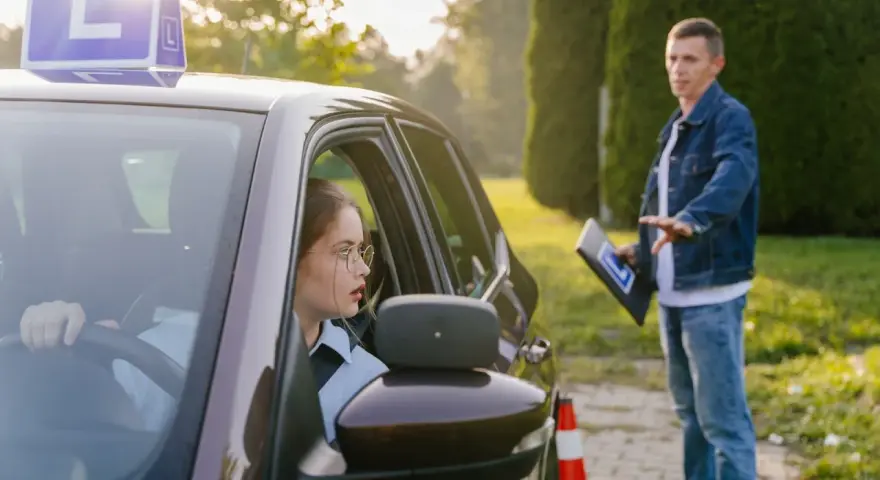 Młoda kobieta w okularach za kierownicą samochodu z literą "L", instruktor obok. Czy kurs na prawo jazdy przepada?