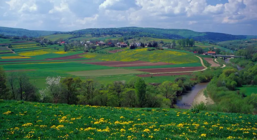 Zielone pogórze z kwitnącymi na żółto łąkami, polami uprawnymi i wijącą się rzeką. W oddali widać zabudowania i lasy.