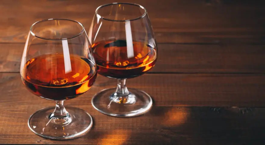 Dwa kieliszki z bursztynowym płynem, przypominającym bourbon lub whiskey, stoją na drewnianym stole.