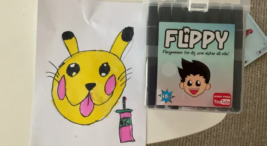 En gul Pikachu-liknande figur med rosa kinder och en tunga som sticker ut, bredvid en låda med färgpennor "FLIPPY" för dig som älskar att rita.