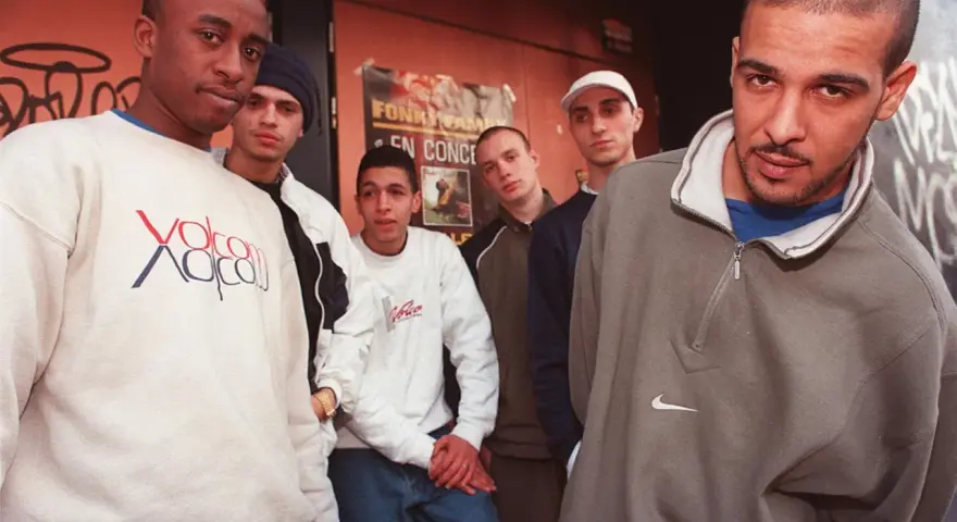 Un groupe de rap français des années 90/2000 pose devant un mur tagué. Ils portent des sweats et des jeans, typiques de l'époque.