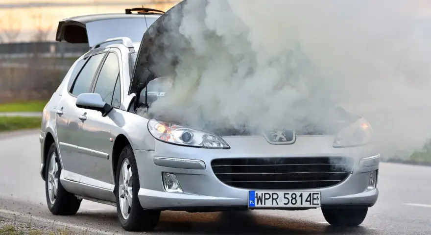 Srebrny samochód Peugeot z otwartą maską, z której wydobywa się gęsty dym.