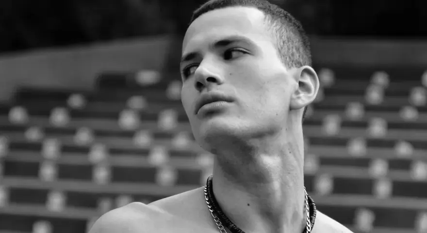 Joven con un corte de pelo buzz cut largo, mirando hacia un lado con una expresión pensativa. Lleva un collar de cadena.