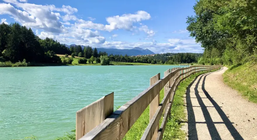 Sentiero sterrato con staccionata in legno lungo il lago di Tavon, circondato da alberi e montagne. Cielo azzurro con nuvole bianche.