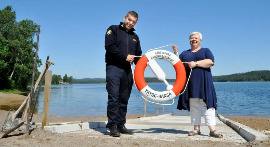 Två personer håller i en räddningsboj vid en sjö. En man i uniform och en kvinna ler. Trygg-Hansa vid Nörybyns camping.