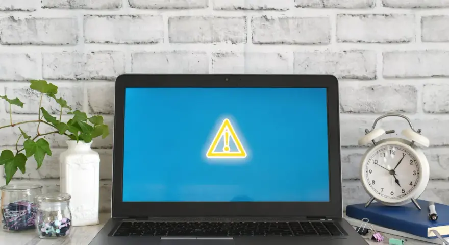 Laptop z symbolem ostrzeżenia na ekranie, obok budzik i rośliny.