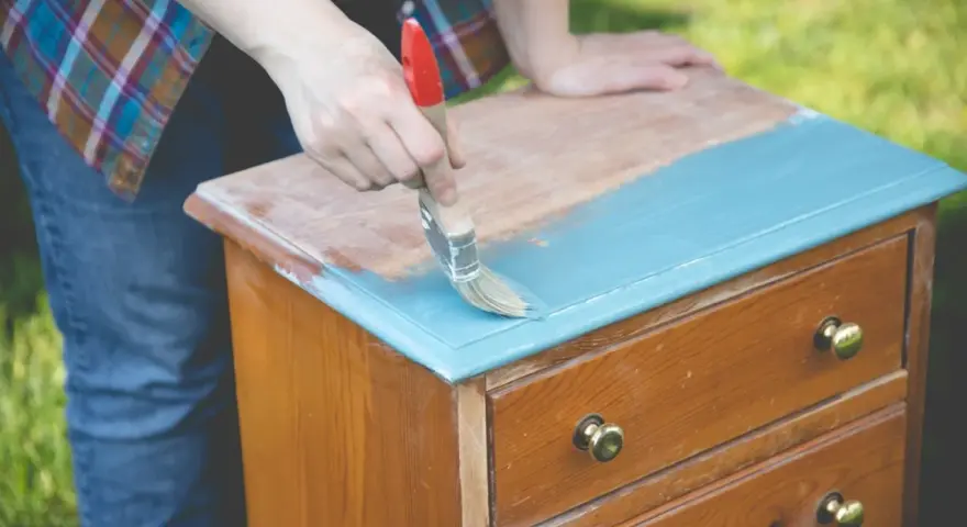 Persona pinta con brocha azul una mesita de noche de madera, transformándola en una de las mesitas de noche pintadas a la tiza.