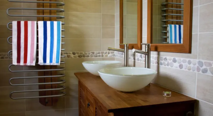 Salle de bain moderne avec double vasque. L'arrivée d'eau est discrète, deux lavabos blancs reposent sur un meuble en bois. Deux serviettes rayées sèchent sur un radiateur.
