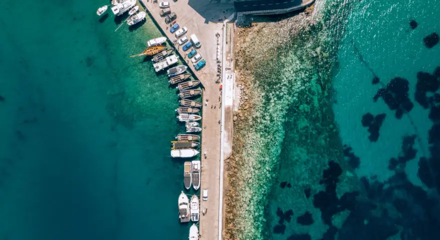 Widok z lotu ptaka na port z licznymi łodziami i jachtami cumującymi przy nabrzeżu. Woda jest krystalicznie czysta, a na dnie widać kamienie i ciemne plamy wodorostów.