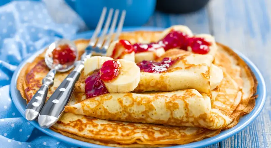 Stos idealnie przygotowanego ciasta na naleśniki, polanego dżemem i kawałkami banana. Obok leżą sztućce.