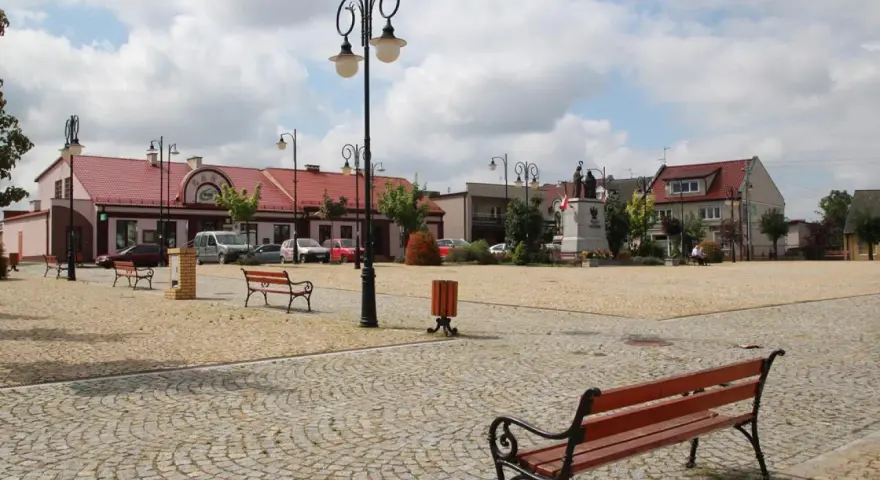 Rynek w Ślesinie z pomnikiem, ławkami i budynkami. Atrakcje tego miejsca zachęcają do odpoczynku.