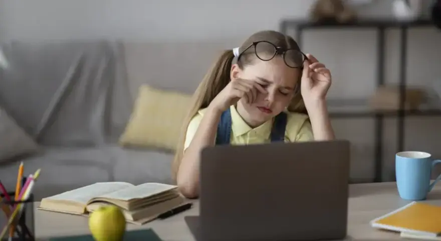 Zmęczona dziewczynka z okularami na czole pociera oczy, ucząc się przy laptopie. Jej indywidualna ścieżka nauczania jest trudna.
