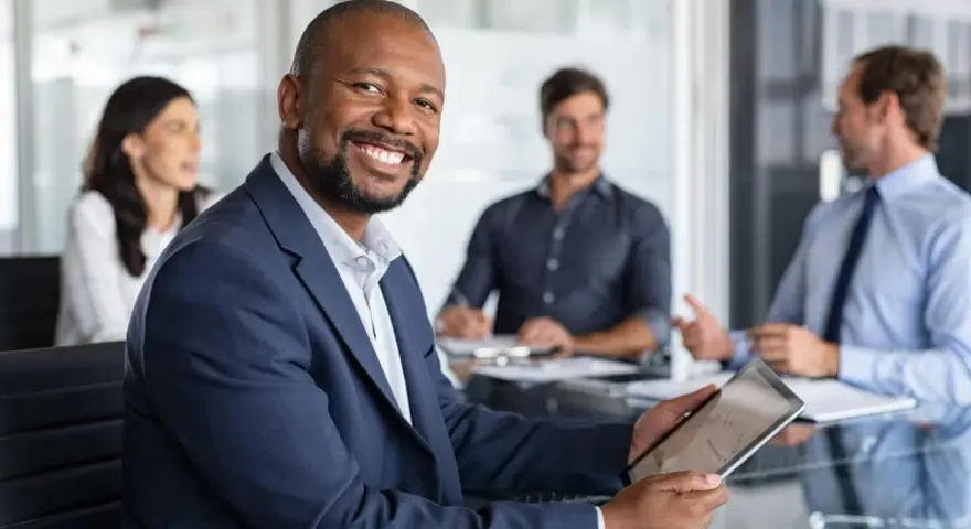 The executive board chair smiles, holding a tablet in a meeting with colleagues.