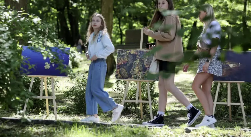 Trzy dziewczyny spacerują wśród obrazów na sztalugach. Jedna z nich, w niebieskich jeansach, przypomina młodszą wersję Tomasza Kłosowskiego.
