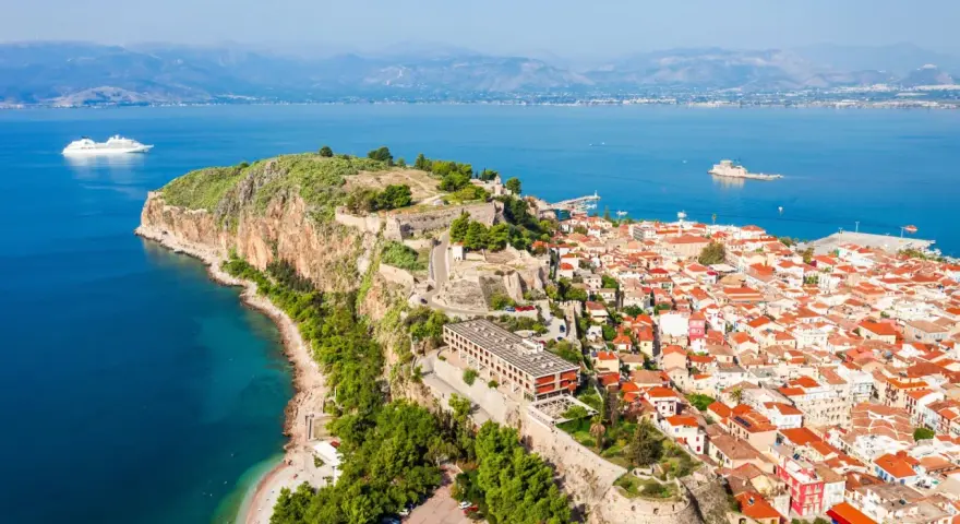 Widok na Nafplio w Grecji: miasto z czerwonymi dachami, twierdza na skale i biały statek wycieczkowy na lazurowym morzu.