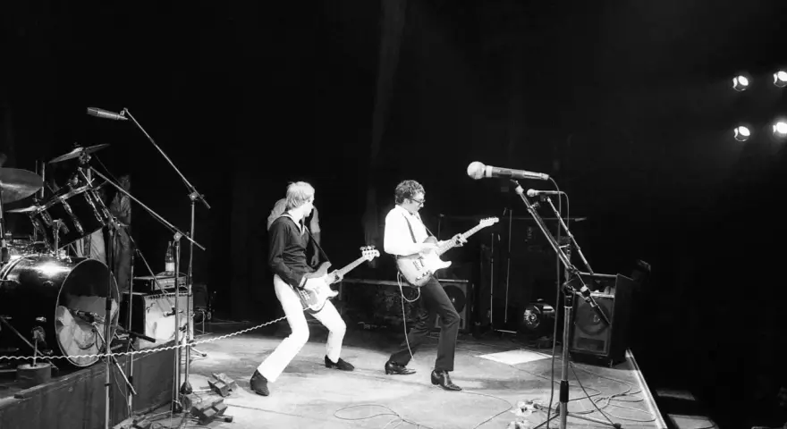 Deux guitaristes d'un groupe français rock jouent sur scène, éclairés par des projecteurs.