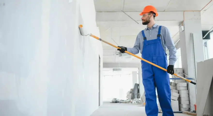 Maler mit blauer Latzhose und orangefarbenem Helm streicht eine Wand weiß. Informieren Sie sich über die malerarbeiten kosten für Ihr Projekt.