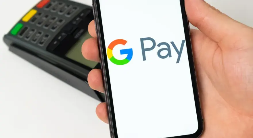 Dłoń trzyma smartfon z widocznym logo Google Pay. W tle terminal płatniczy. Szybkie i wygodne płatności.