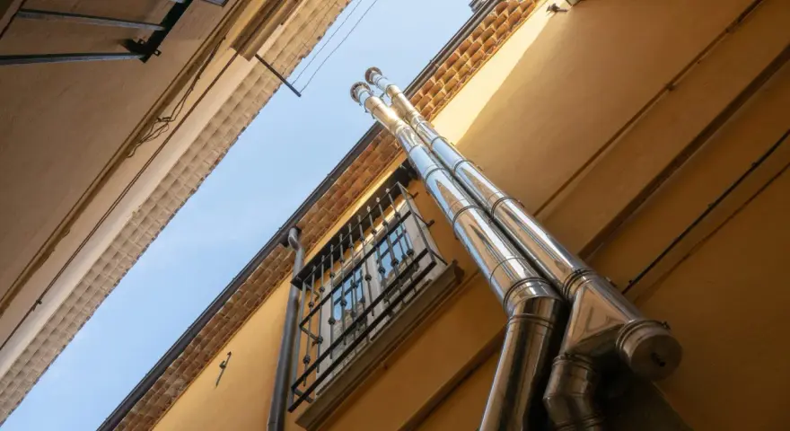 Dos tubos de salida humos estufa pellets brillan bajo el sol, ascendiendo por la pared amarilla de un edificio.
