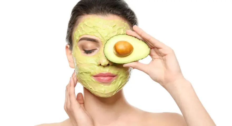 Frau mit Avocado-Gesichtsmaske und Avocado-Scheibe vor dem Auge. Die Avocado-Wirkung auf die Haut ist bekannt für ihre pflegenden Eigenschaften.